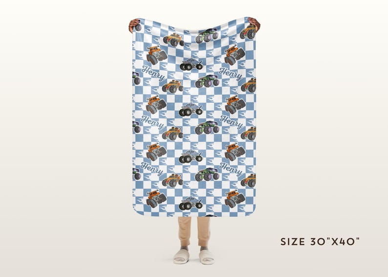 May include: A blue and white chequered baby blanket with a repeated pattern of monster trucks. The trucks are orange, black, and grey. The blanket has the name "Henry" printed on each truck. The blanket is 76 cm by 102 cm.