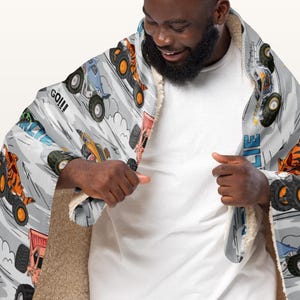 May include: A hooded blanket with a monster truck design on a light grey background. The design includes various monster trucks in action, with the word "GO!!!" visible. The blanket has a soft, plush interior.