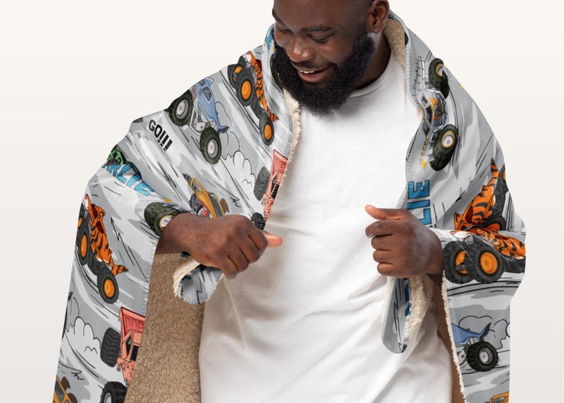 May include: A hooded blanket with a monster truck design on a light grey background. The design includes various monster trucks in action, with the word "GO!!!" visible. The blanket has a soft, plush interior.