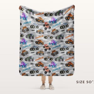 May include: A grey blanket featuring a pattern of colourful monster trucks, some with the name "Charlie" and the word "GO!!!". The blanket measures 127cm x 152cm.