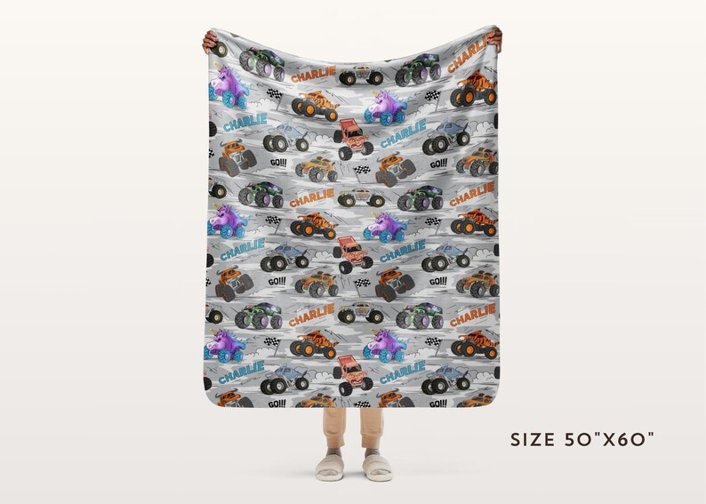 May include: A grey blanket featuring a pattern of colourful monster trucks, some with the name "Charlie" and the word "GO!!!". The blanket measures 127cm x 152cm.