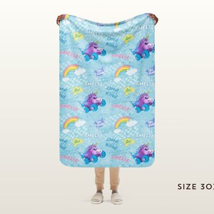 May include: A blue fleece blanket with a repeating pattern of a unicorn riding a skateboard, rainbows, clouds, and the text "Amelia" and "One Kind". The blanket is 76 cm by 102 cm.