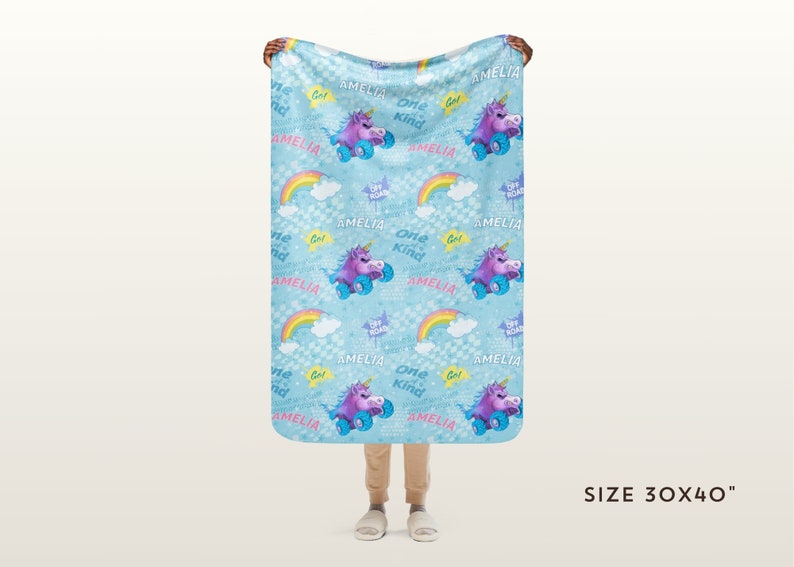 May include: A blue fleece blanket with a repeating pattern of a unicorn riding a skateboard, rainbows, clouds, and the text "Amelia" and "One Kind". The blanket is 76 cm by 102 cm.