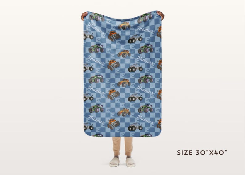 May include: A blue and white checkered fleece blanket with a repeating pattern of monster trucks and the name "Thomas". The blanket is 30 inches by 40 inches.