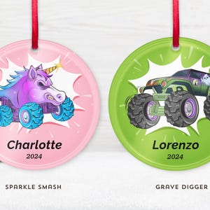 May include: Two round Christmas ornaments with red ribbons. One pink ornament features a cartoon unicorn monster truck with the name "Charlotte" and the year "2024". The other green ornament shows a monster truck with the name "Lorenzo" and the year "2024".