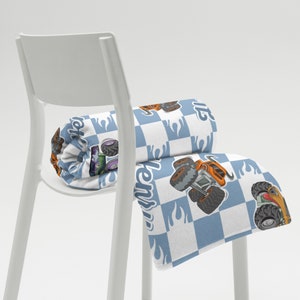 May include: A white and blue checkered throw blanket with a monster truck design. The blanket is folded and placed on a white chair. The blanket has the word "monster" printed on it.