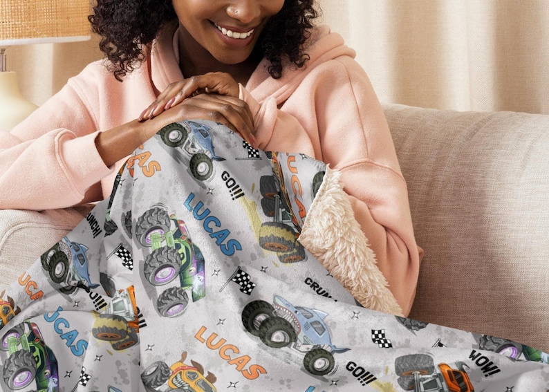 Op de afbeelding: Een grijs en wit fleece deken met een herhalend patroon van monstertrucks. De trucks zijn blauw, groen, oranje en geel. Het deken heeft de naam "Lucas" meerdere keren erop gedrukt. Het deken wordt vastgehouden door een persoon die op een bank zit.
