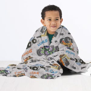 Op de afbeelding: Een grijs en wit fleece deken met een monstertruck thema. Het deken heeft afbeeldingen van monstertrucks, de woorden "Lucas", "Go!!!", en "Monster Truck" in zwart en wit.