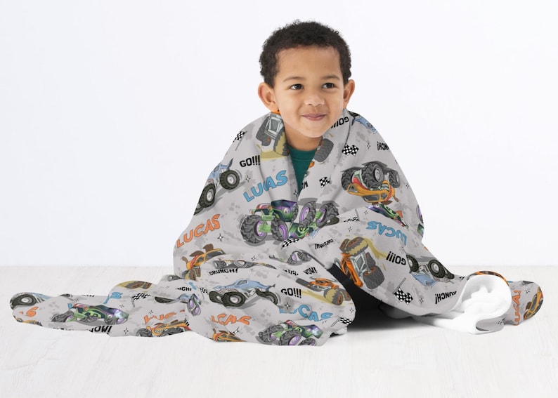 Op de afbeelding: Een grijs en wit fleece deken met een monstertruck thema. Het deken heeft afbeeldingen van monstertrucks, de woorden "Lucas", "Go!!!", en "Monster Truck" in zwart en wit.