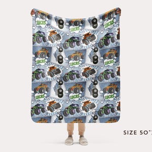Puede incluir: Una manta de franela azul y blanca con un patr&oacute;n repetido de monster trucks. Los camiones son verdes, naranjas y negros con el nombre "Lucas" en un bocadillo. La manta mide 127 cm por 152 cm.