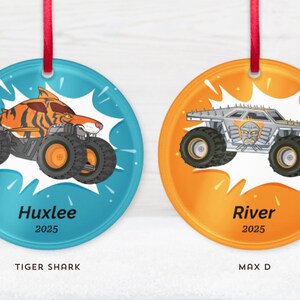 May include: Two round Christmas ornaments with red ribbons. One features a tiger shark monster truck on a blue background, the other a silver monster truck on an orange background. The names "Huxlee" and "River" are printed on the ornaments.