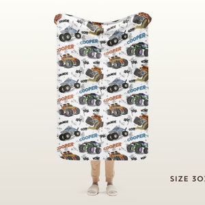 May include: A white fleece blanket with a repeating pattern of monster trucks in various colours, including orange, blue, and green. The trucks are driving on a dirt track and have the word "COOPER" printed on them. The blanket is 76 cm by 102 cm.