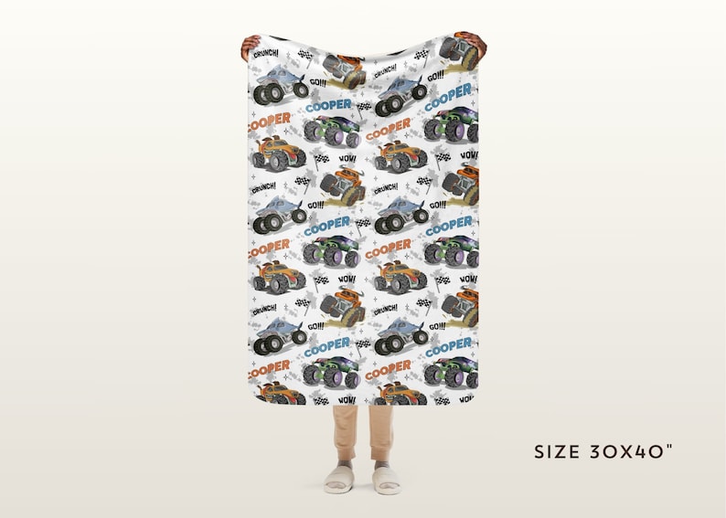 May include: A white fleece blanket with a repeating pattern of monster trucks in various colours, including orange, blue, and green. The trucks are driving on a dirt track and have the word "COOPER" printed on them. The blanket is 76 cm by 102 cm.