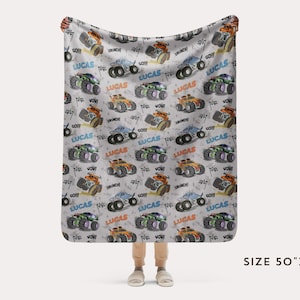 Op de afbeelding: Een wit fleece deken met een herhalend patroon van monstertrucks in verschillende kleuren. De trucks hebben de naam "Lucas" erop gedrukt. Het deken is 50 inch bij 60 inch.