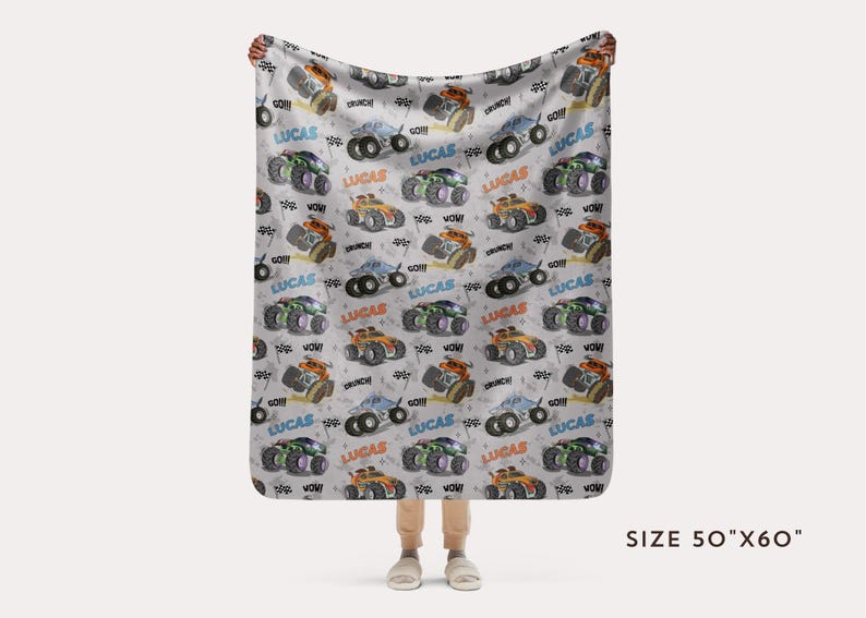 Op de afbeelding: Een wit fleece deken met een herhalend patroon van monstertrucks in verschillende kleuren. De trucks hebben de naam "Lucas" erop gedrukt. Het deken is 50 inch bij 60 inch.