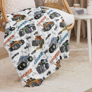 May include: White fleece blanket featuring a repeating pattern of monster trucks in various colours. The trucks have the name "Cooper" written on them and phrases like "Go!" and "Crunch!" The blanket is ideal for a child's room.