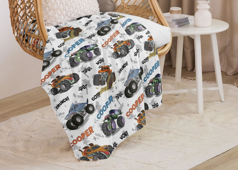 May include: White fleece blanket featuring a repeating pattern of monster trucks in various colours. The trucks have the name "Cooper" written on them and phrases like "Go!" and "Crunch!" The blanket is ideal for a child's room.