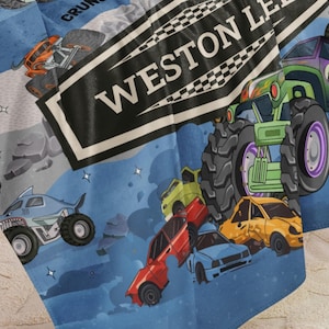 May include: A blue blanket featuring a colorful monster truck design. The blanket has illustrations of monster trucks, crushed cars, and the text "WESTON LEAGUE". The design includes a shark-themed monster truck and a green monster truck.