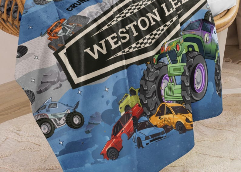 May include: A blue blanket featuring a colorful monster truck design. The blanket has illustrations of monster trucks, crushed cars, and the text "WESTON LEAGUE". The design includes a shark-themed monster truck and a green monster truck.