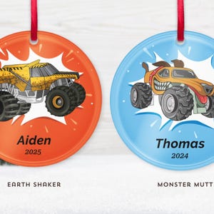 May include: Two round Christmas ornaments with red ribbons. One is orange with a yellow monster truck and the name "Aiden" and the year "2025". The other is blue with a monster truck and the name "Thomas" and the year "2024".