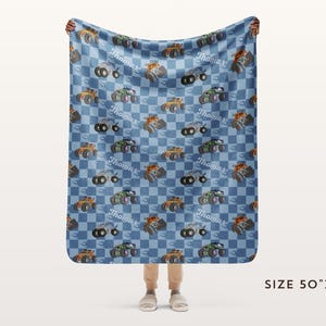 May include: A blue and white checkered fleece blanket with a repeated pattern of monster trucks. The trucks are orange, black, and gray with white wheels. The blanket is personalized with the name "Thomas" repeated throughout the pattern. The blanket is 50 inches by 60 inches.