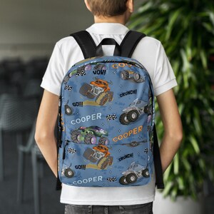 May include: Blue backpack with a monster truck pattern. The backpack features the words "COOPER", "GO!!!", "WOW!" and "CRUNCH!" repeated throughout the design.