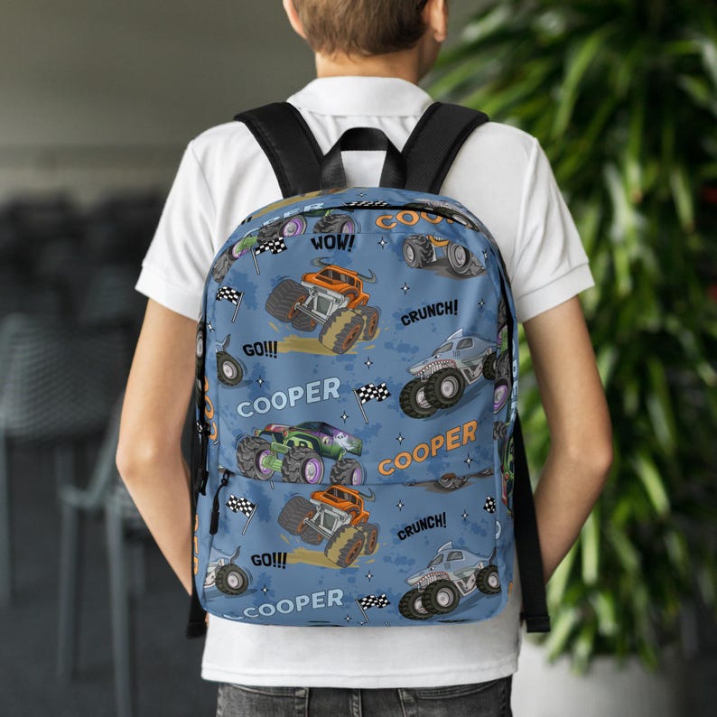May include: Blue backpack with a monster truck pattern. The backpack features the words "COOPER", "GO!!!", "WOW!" and "CRUNCH!" repeated throughout the design.