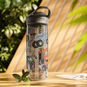 May include: Clear water bottle with a black lid and handle, featuring monster truck graphics in blue, green, and orange. The bottle has the name "JAMES" printed on it, along with the word "WOW!"
