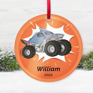 May include: Orange round ornament with a cartoon shark monster truck design. The shark truck is light blue with large black tires. The ornament has the name "William" and the year "2024" printed on it.