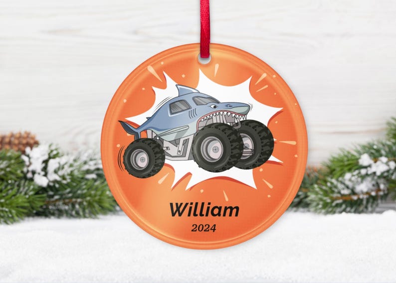 May include: Orange round ornament with a cartoon shark monster truck design. The shark truck is light blue with large black tires. The ornament has the name "William" and the year "2024" printed on it.