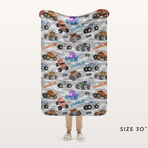 May include: A grey blanket featuring a pattern of colourful monster trucks, a unicorn, and the name "Charlie". The blanket measures 76cm x 102cm.