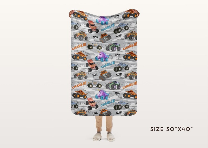 May include: A grey blanket featuring a pattern of colourful monster trucks, a unicorn, and the name "Charlie". The blanket measures 76cm x 102cm.