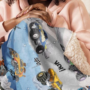 May include: A blue, gray, and white monster truck blanket with a soft, plush sherpa lining. The blanket features colorful monster truck illustrations, a checkered flag, and the word "WOW!"