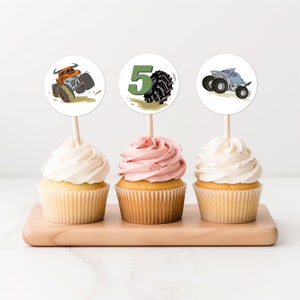 May include: Three cupcakes with monster truck themed toppers. The cupcakes are frosted with white and pink frosting. The toppers are round and feature a monster truck, a number 5, and a monster truck with a shark fin.