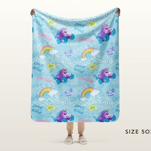 May include: A blue fleece blanket with a repeating pattern of rainbows, clouds, unicorns riding off-road vehicles, and the text "Amelia" and "One Kind Girl". The blanket is 127 cm by 152 cm.