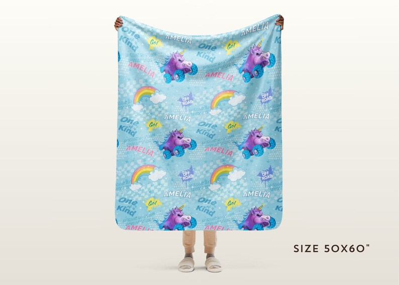 May include: A blue fleece blanket with a repeating pattern of rainbows, clouds, unicorns riding off-road vehicles, and the text "Amelia" and "One Kind Girl". The blanket is 127 cm by 152 cm.
