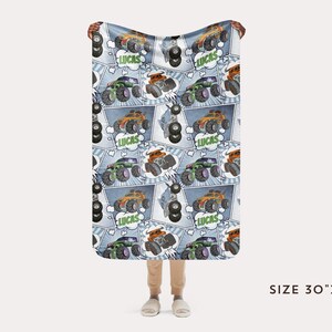 Puede incluir: Una manta de franela azul y blanca con un patr&oacute;n repetido de monster trucks, neum&aacute;ticos y la palabra "Lucas". La manta mide 76 cm por 102 cm.