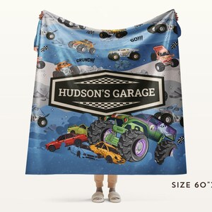 May include: A blue blanket featuring a colorful monster truck design with the text "HUDSON'S GARAGE". The blanket has a size of 60" x 80" and includes illustrations of monster trucks and race flags. The design includes the words "Crunch!" and "GO!!!".
