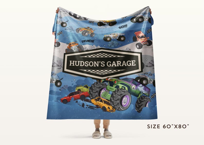 May include: A blue blanket featuring a colorful monster truck design with the text "HUDSON'S GARAGE". The blanket has a size of 60" x 80" and includes illustrations of monster trucks and race flags. The design includes the words "Crunch!" and "GO!!!".