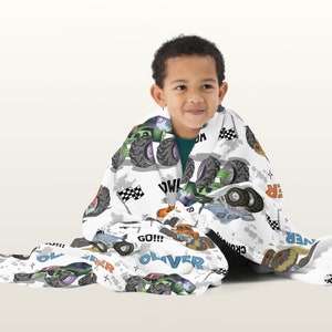 May include: A white fleece blanket with a monster truck theme. The blanket features a variety of monster trucks in different colours, including green, orange, blue, and black. The blanket also features the words "GO!!!", "WOW!" and "OLIVER" printed on it.