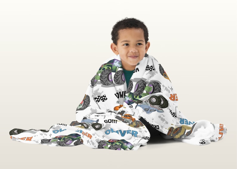 May include: A white fleece blanket with a monster truck theme. The blanket features a variety of monster trucks in different colours, including green, orange, blue, and black. The blanket also features the words "GO!!!", "WOW!" and "OLIVER" printed on it.