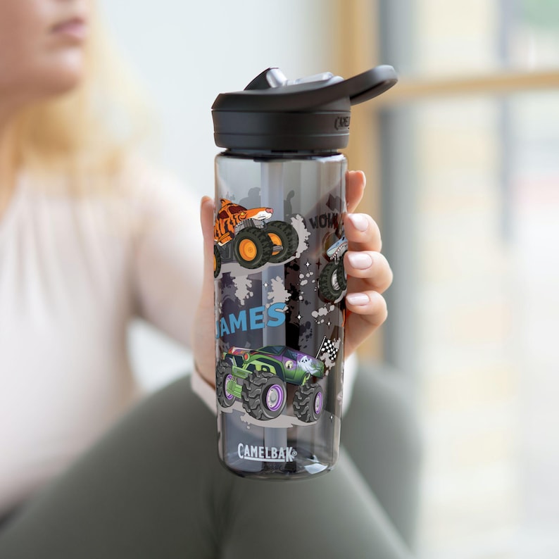 May include: A clear water bottle with a black lid and a flip-top spout. The bottle features colorful monster truck graphics and the word "GAMES" in blue. The brand name "CAMELBAK" is printed at the bottom.
