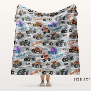 May include: A grey blanket featuring a pattern of colourful cartoon monster trucks. The blanket has the name "Charlie" and the word "GO!!!" printed on it. The blanket measures 152cm x 203cm.