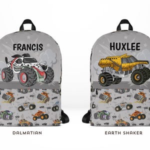 Może przedstawiać: Dwa szare plecaki z czarnymi paskami. Jeden ma wz&oacute;r monster trucka dalmatyńczyka z napisem "FRANCIS" powyżej. Drugi ma wz&oacute;r monster trucka Earth Shaker z napisem "HUXLEE". Oba mają powtarzający się wz&oacute;r monster trucka.