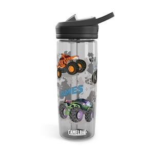 May include: Clear water bottle with a black lid and straw. The bottle features colorful monster truck graphics, including a tiger-themed truck and the name "JAMES". The brand name "CAMELBAK" is printed at the bottom.