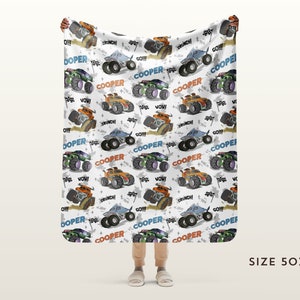 May include: A white fleece blanket featuring a repeating pattern of monster trucks in various colours, including orange, blue, and green. The trucks have large wheels and are driving on a white background with black tyre tracks. The words "COOPER", "GO!!!", "WOW", "CRUNCH", and "O!!!" are printed on the blanket.