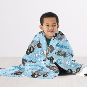 Pu&ograve; includere: Una coperta in pile blu e grigia con un design di camion mostro. La coperta presenta il nome "Cooper" e la frase "Off Road" ripetuta su tutta la superficie. Un bambino &egrave; seduto sul pavimento con la coperta avvolta intorno a lui.