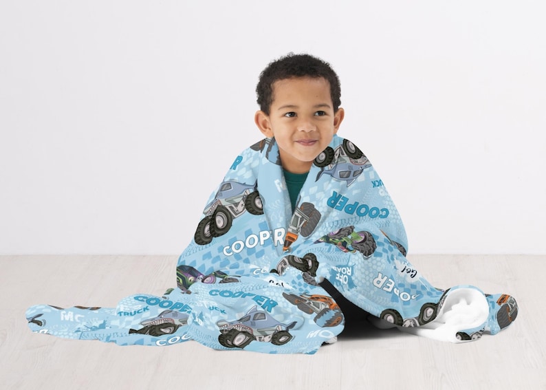 Pu&ograve; includere: Una coperta in pile blu e grigia con un design di camion mostro. La coperta presenta il nome "Cooper" e la frase "Off Road" ripetuta su tutta la superficie. Un bambino &egrave; seduto sul pavimento con la coperta avvolta intorno a lui.