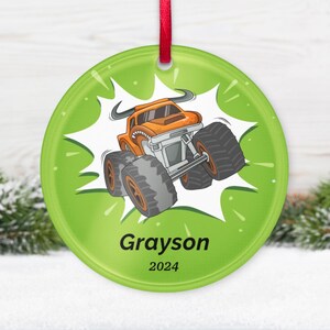 May include: A round, green Christmas ornament with a cartoon monster truck design. The orange truck has large tires and is set against a white burst. The name "Grayson" and the year "2024" are printed below the truck. A red ribbon is attached for hanging.