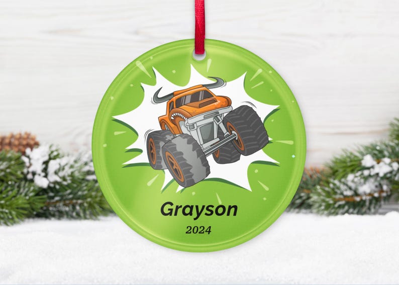 May include: A round, green Christmas ornament with a cartoon monster truck design. The orange truck has large tires and is set against a white burst. The name "Grayson" and the year "2024" are printed below the truck. A red ribbon is attached for hanging.